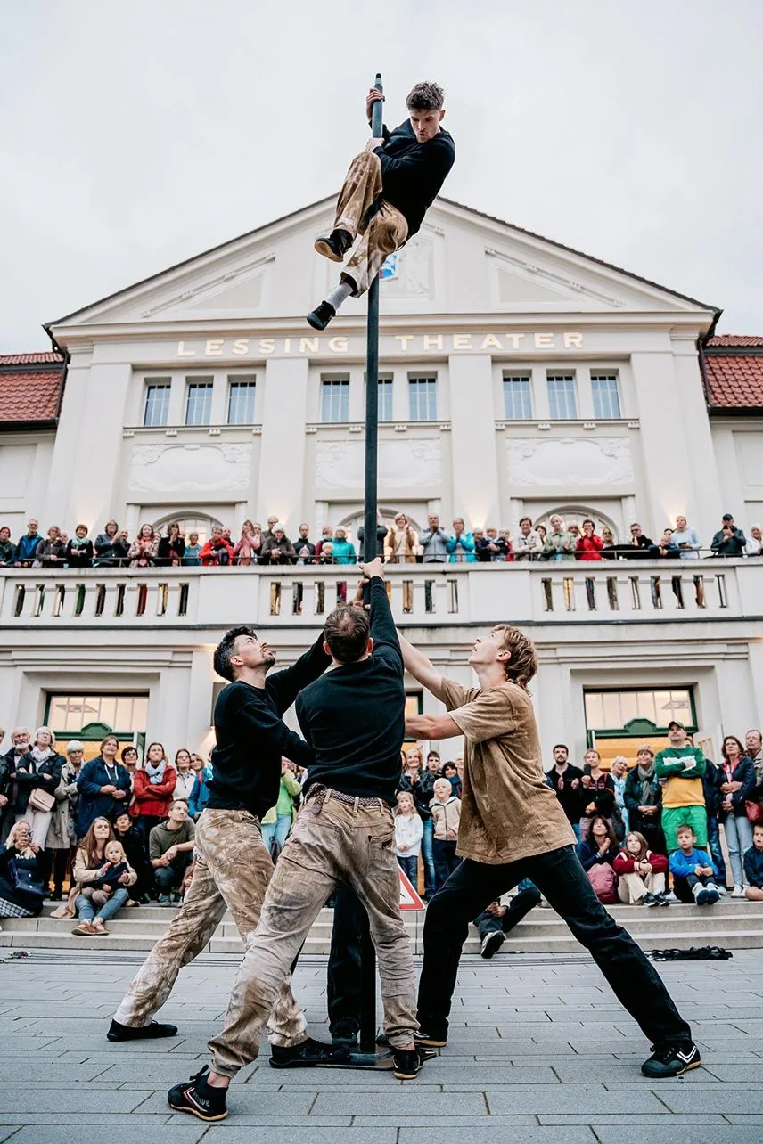 Ein Akrobat hält sich hoch oben am Mast, den mehrere Kollegen am Boden stützen. Zuschauer stehen vor dem Lessingtheater und auf dessen Terrasse.