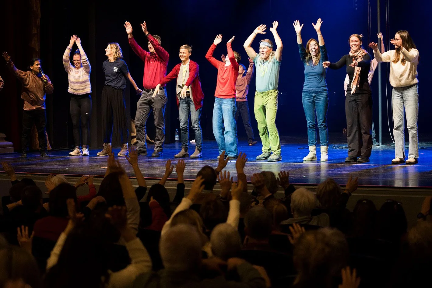 Eine Reihe Menschen auf der Theaterbühne hebt die Arme und klatscht. Im Zuschauerraum strecken Gäste ebenfalls die Hände nach oben.